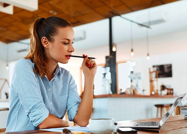 woman with pencil in her mouth, thinking hard as she writes a blog on computer