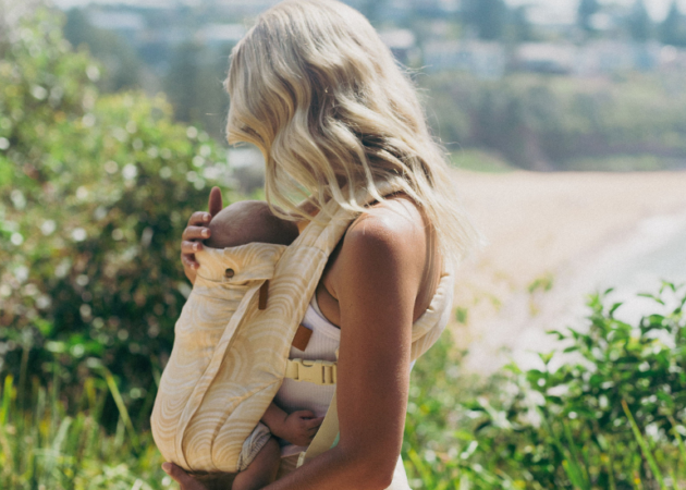 women holding baby with Sia baby carrier, backdrop is of greenery overlooking a beach