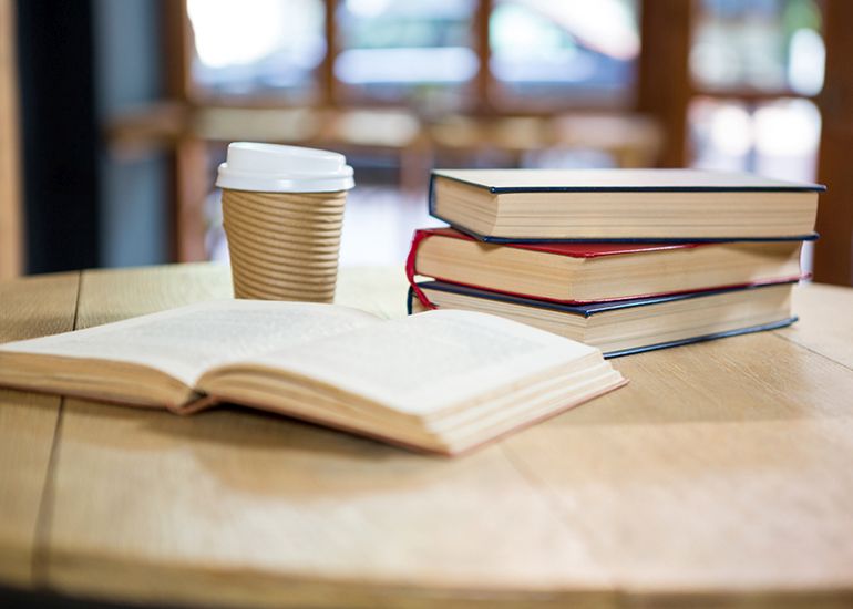 Book lying open on table beside a pile of books and a takeaway coffee cup