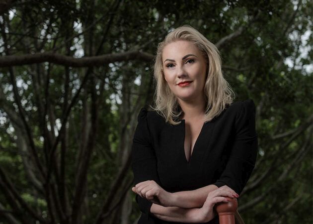 Taminda Polle headshot, blonde woman in a black dress stands in front of a fig tree.