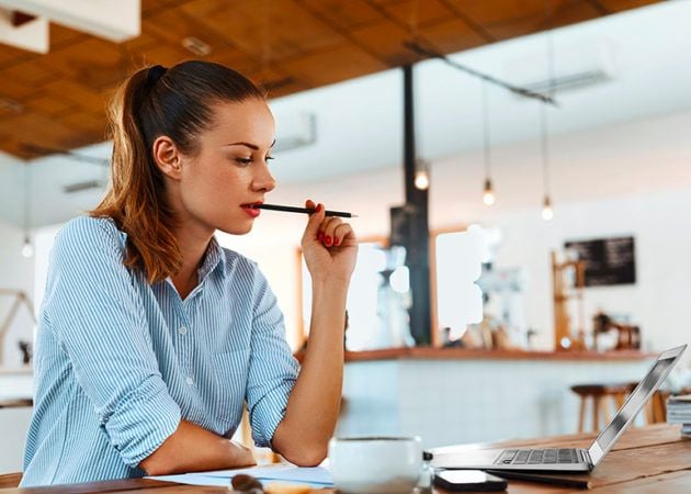 woman with pencil in her mouth, thinking hard as she writes a blog on computer