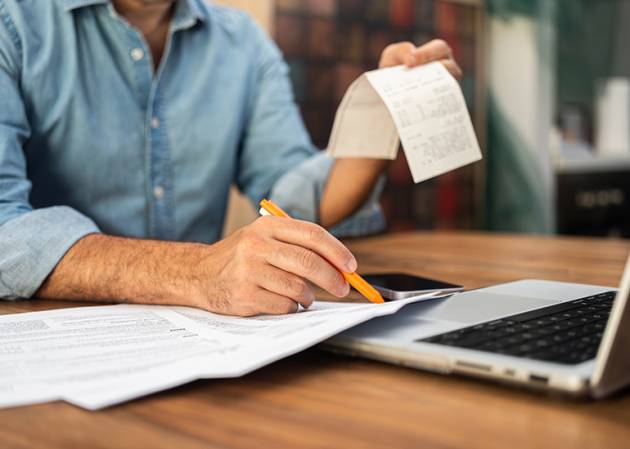 Man filling out tax forms at home using a laptop and holding receipts