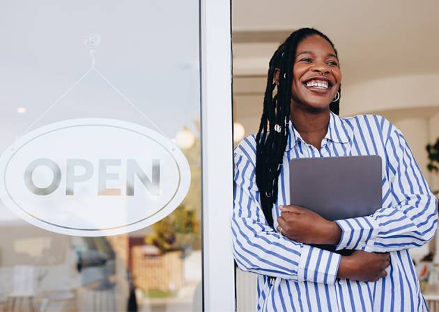 A confident smiling business owner stands in the door of a shop holding a laptop to her chest.