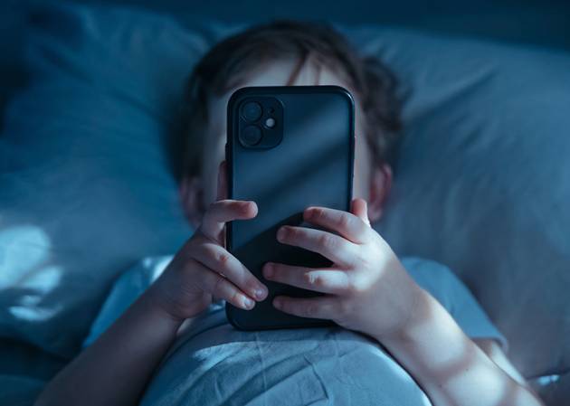 Close-up of unrecognisable little child lying in bed at night, holding mobile phone in front of his face. Gadget addiction and excessive late-night screen time stealing life